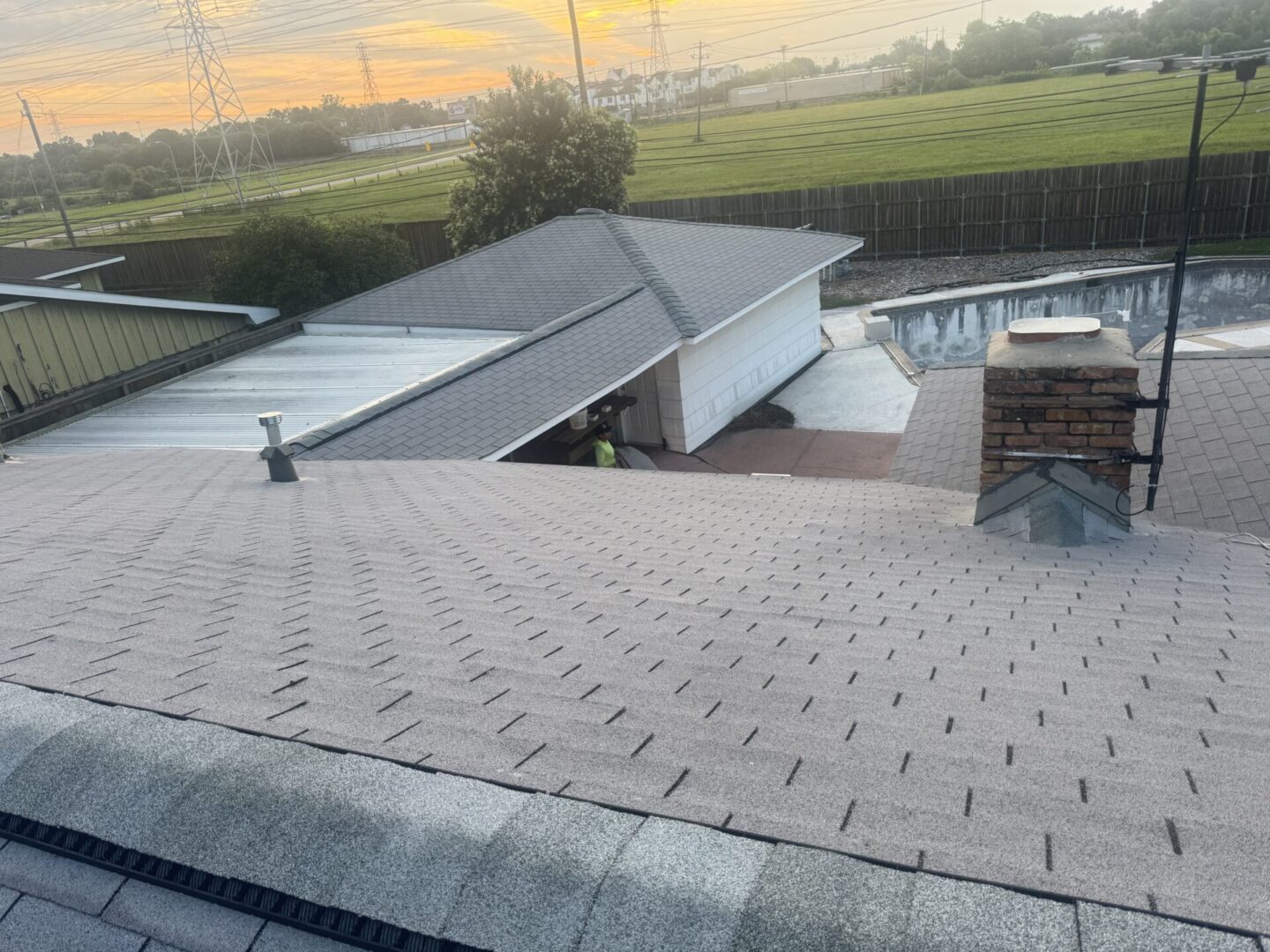 An elevated view from a rooftop showcases newly installed shingles and a brick chimney overlooking a detached garage and backyard pool during a golden sunset, emphasizing the value of timely and affordable roof repair.