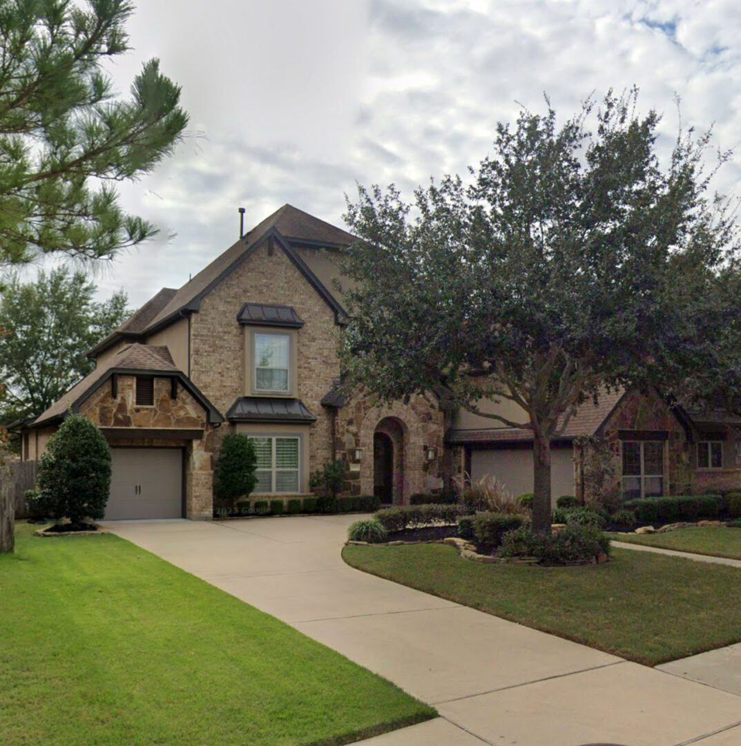 A large, multi-level home featuring an elegant mix of brick and stone siding stands behind a beautifully landscaped front lawn and paved driveway, showcasing a pristine residential roof replacement.