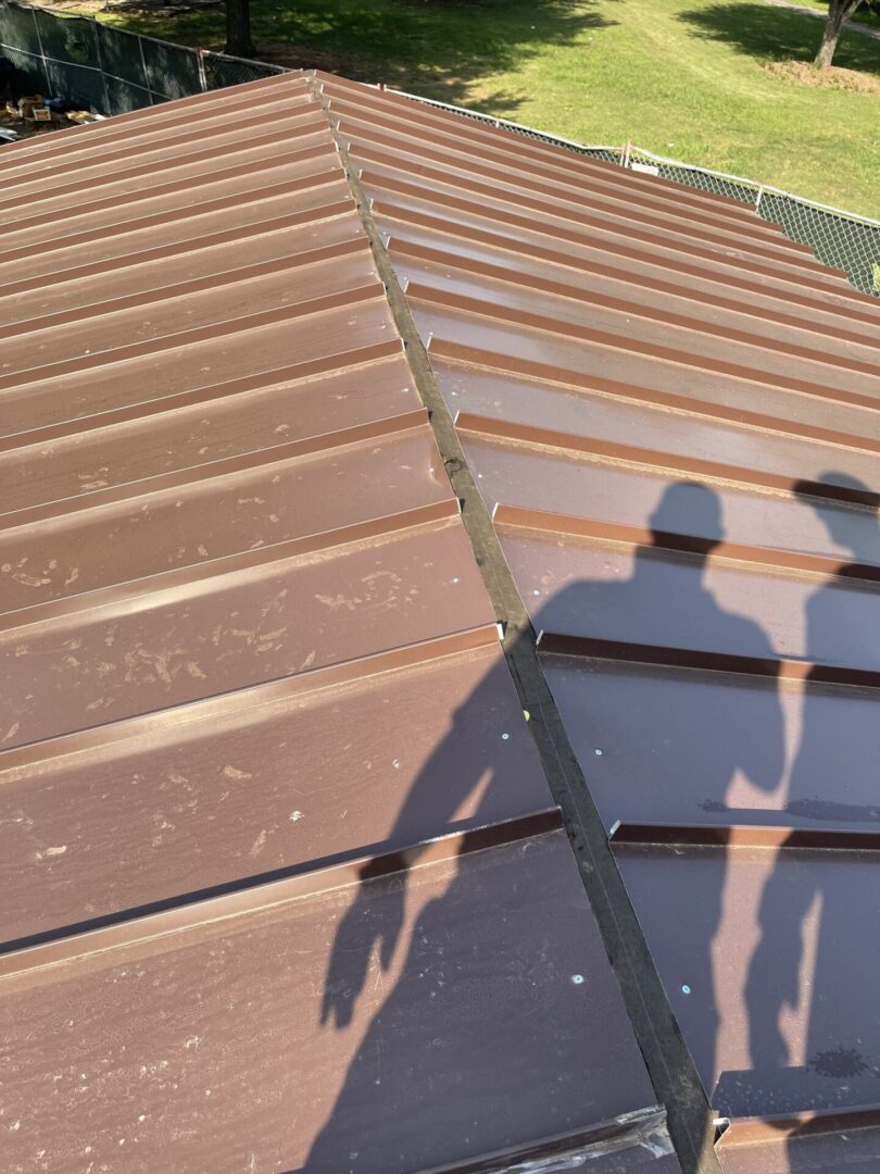 A brown standing-seam metal roof installation highlights a residential roof replacement project on a clear day, with a view of the surrounding green lawn and chain-link fence.