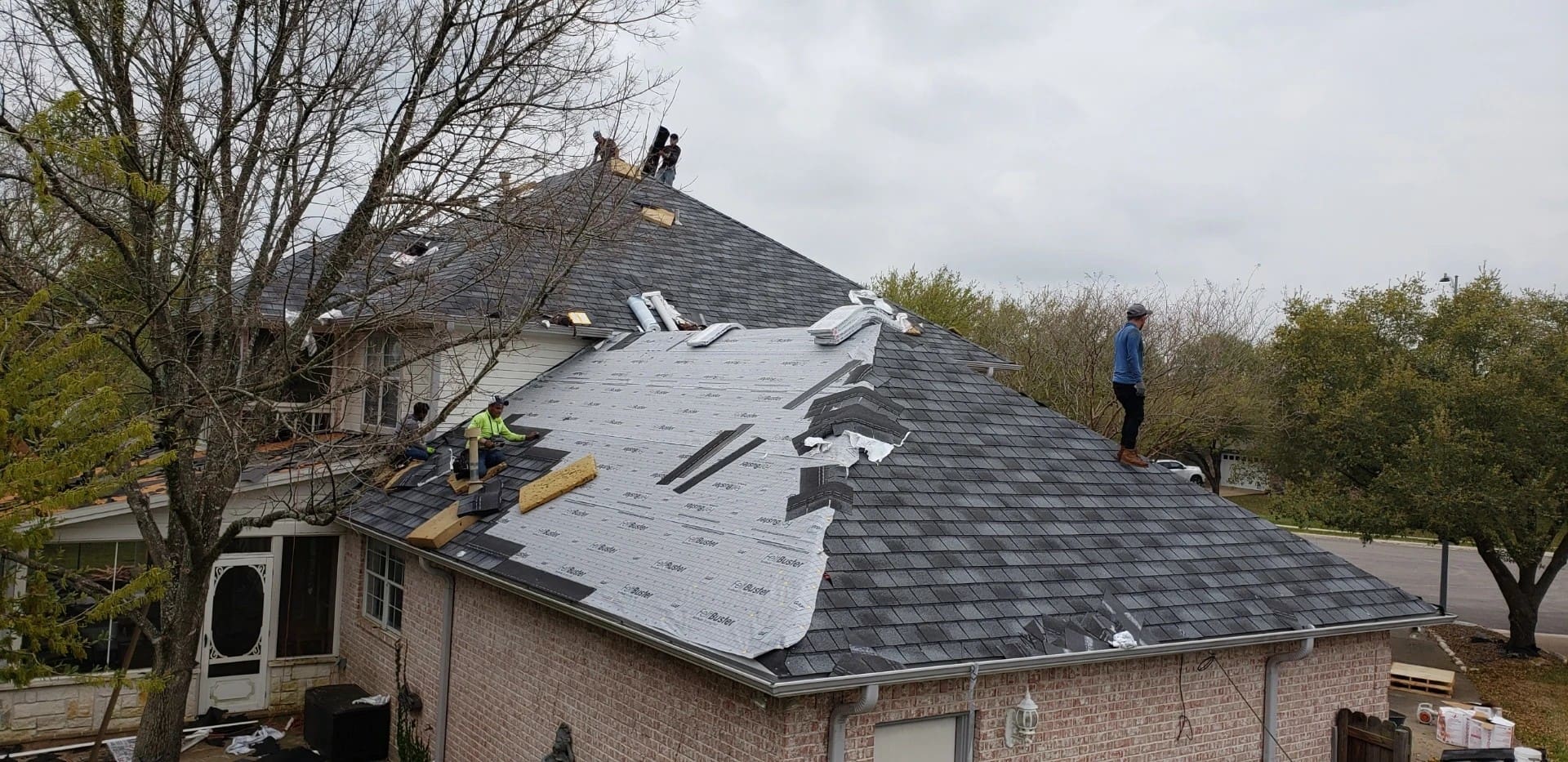 Several workers are actively installing new shingles and protective underlayment on a multi-level home during a residential roof replacement.