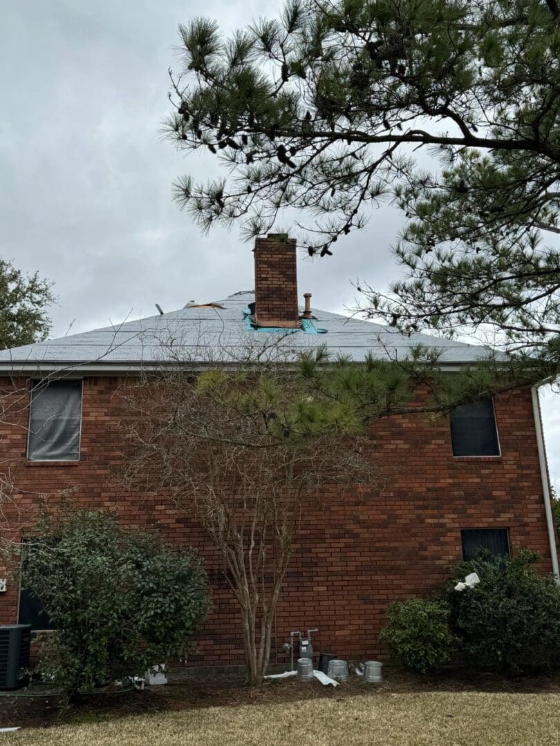 A two-story brick home stands with its upper shingles removed and an underlayment layer exposed in preparation for a residential roof repair.